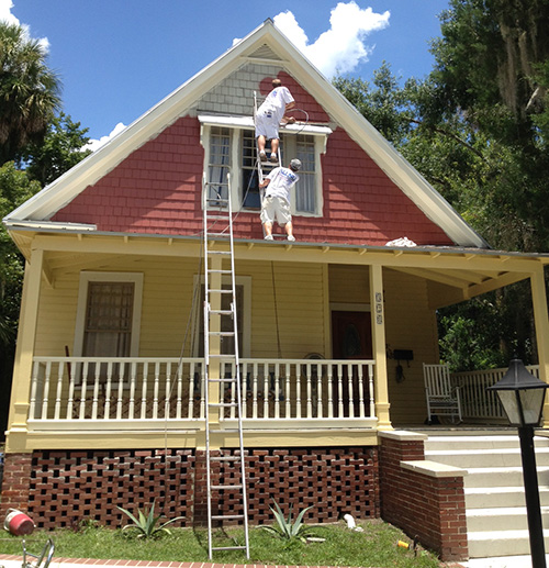 Red house with ladder and painters on roof.